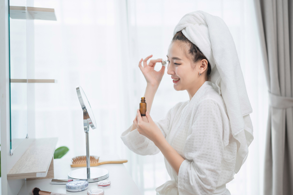 Woman with towel on head applying skincare serum in bright bathroom setting, enjoying self-care routine and beauty therapy at home