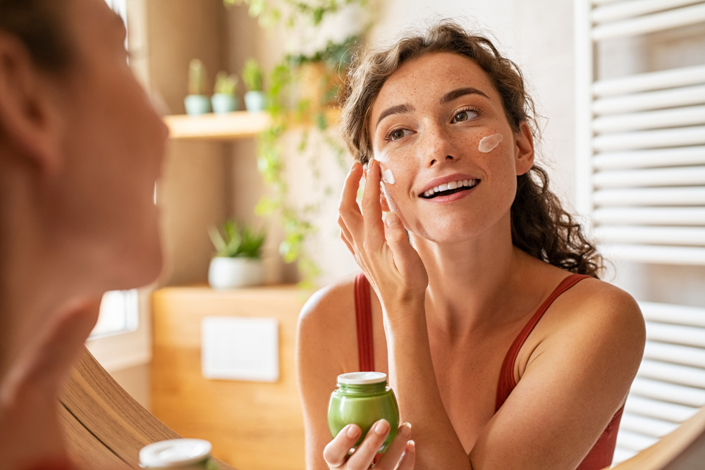 Woman caring of her beautiful skin face standing near mirror in the bathroom. Young woman applying moisturizing cream on her face during morning routine