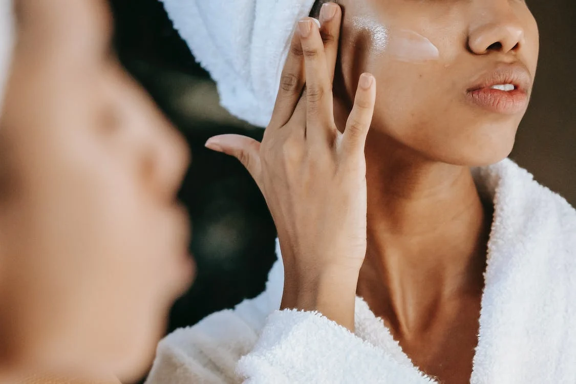 crop ethnic woman applying cream on the face against the mirror